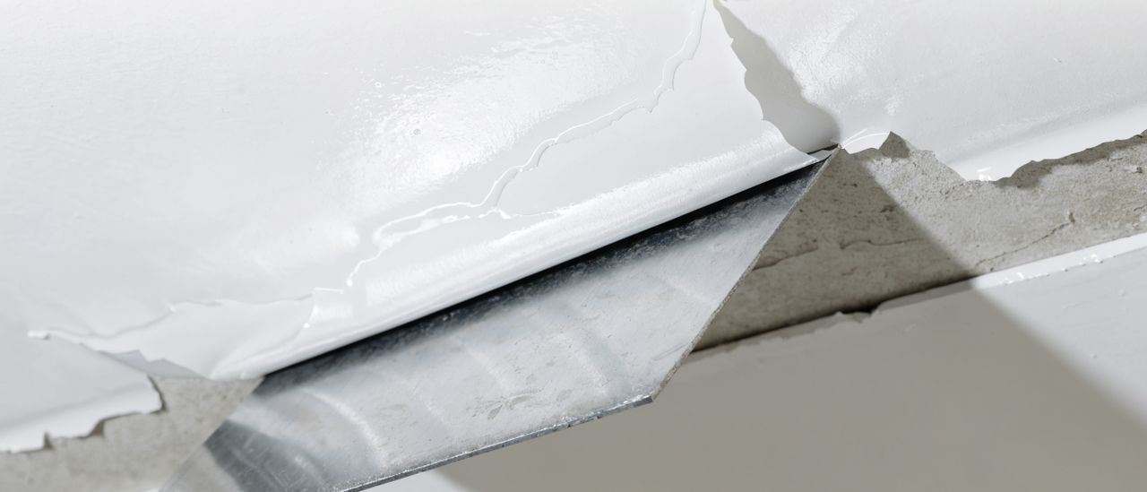 Close-up of a scraper removing wet popcorn ceiling texture from drywall